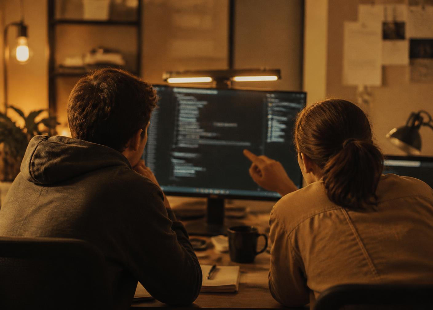 Two engineers reviewing code together on a large monitor in a warmly lit workspace, seen from behind